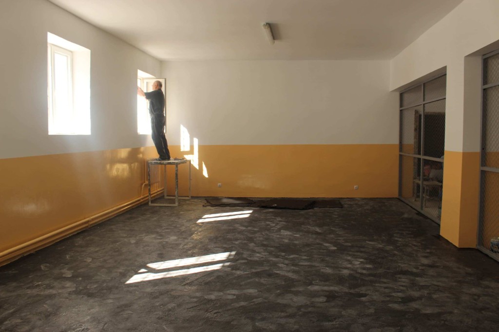 A prisoner painting a window in the newly plastered gymnasium, with yellow lower walls and a finished floor