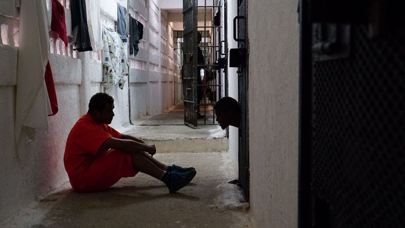 Two people in a prison hallway, one sitting on the floor and another looking through a cell door hatch, illustrating a human connection.