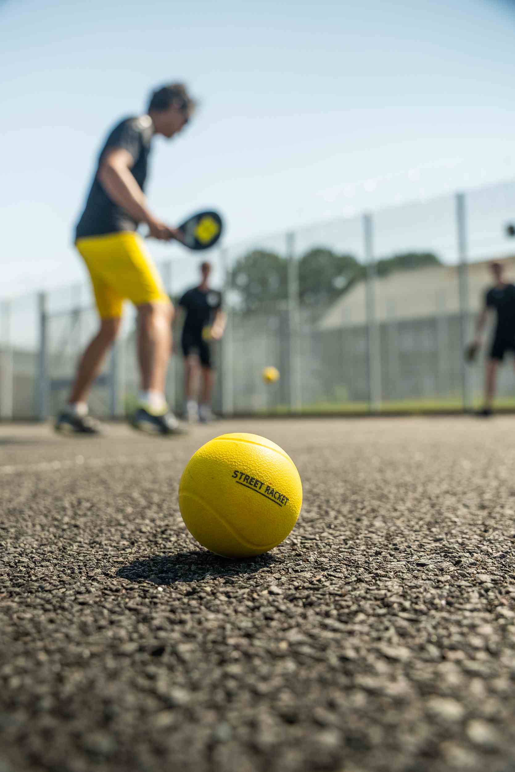 Close-up of a yellow Street Racket ball with players in the background